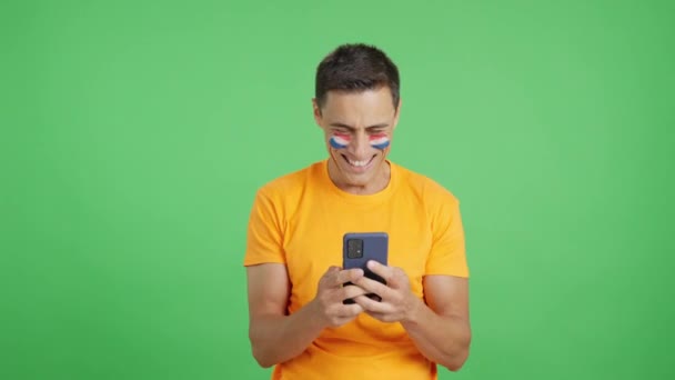 Studio video s chromatickým pozadím holandského fanouška s vlajkou Holandska namalovanou na tváři, dívá se na svůj mobilní telefon s úsměvem a ukazuje ho kameře s obrazovkou v chromatickém klíči - Záběry, video