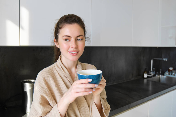 Pojęcie stylu życia. Portret szczęśliwej brunetki w szlafroku, pijącej kawę w kuchni, pijącej poranną herbatę i uśmiechającej się. - Zdjęcie, obraz