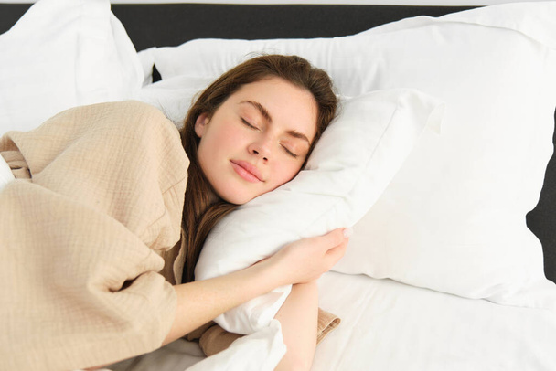 Porträt einer glücklichen, schönen jungen Frau, die im Bett liegt, Schlafanzug trägt, ihr Kopfkissen umarmt und im Schlaf lächelt. - Foto, Bild