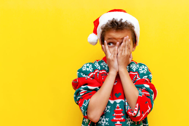 ντροπαλό αφροαμερικανό αγόρι με χριστουγεννιάτικα ρούχα καλύπτει το πρόσωπό του με τα χέρια του και κρυφοκοιτάζει σε κίτρινο απομονωμένο φόντο, φοβισμένο 9χρονο παιδί με καπέλο Άι Βασίλη κατασκοπεύει - Φωτογραφία, εικόνα
