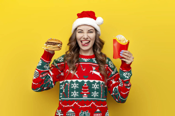 молода мила дівчина в різдвяному светрі і капелюсі Санта Клауса, що тримає бургер і картоплю фрі і показує язик на жовтому ізольованому фоні, жінка їсть фаст-фуду на новий рік - Фото, зображення