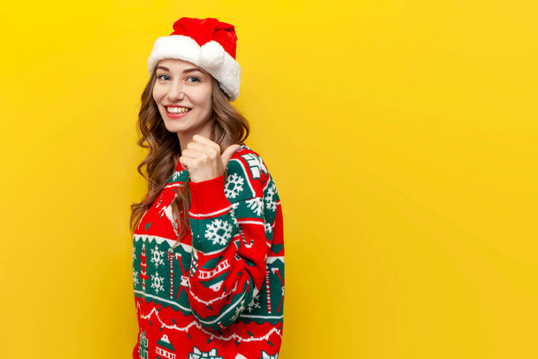 jeune fille en pull de Noël et santa claus chapeau sourit et pointe vers l'arrière sur fond jaune isolé, femme en vêtements de Noël pointe avec ses mains à l'espace de copie par derrière - Photo, image