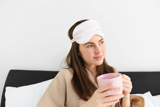 Wspaniała kobieta w piżamie i masce sypialnej, trzymająca filiżankę kawy, pijąca cappuccino w łóżku i wyglądająca na szczęśliwą, odpoczywająca w sypialni w weekend, mająca relaksujący poranek. - Zdjęcie, obraz