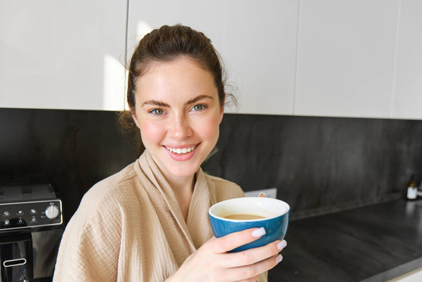 Przystojna młoda kobieta z filiżanką kawy, pozująca w kuchni, uśmiechnięta do kamery, cieszy się porankiem w domu, ubrana w przytulny szlafrok. - Zdjęcie, obraz