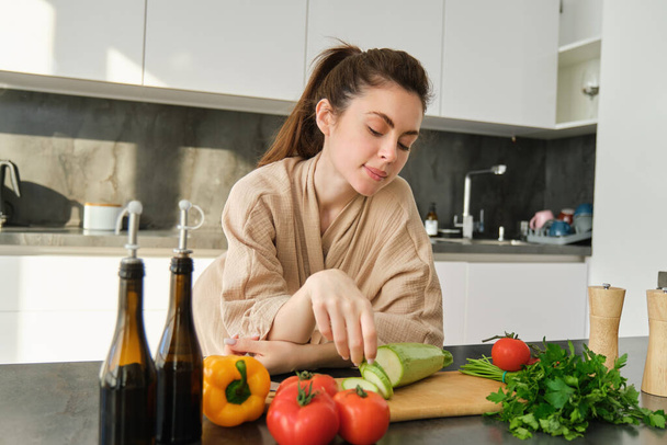 Портрет милой молодой женщины в халате, приготовление пищи, стоя рядом с доской с овощами, проведение цуккини, приготовление салата или вегетарианский ужин. - Фото, изображение