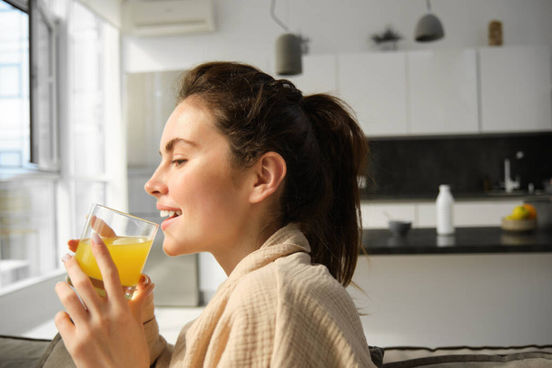 Крупним планом портрет щасливої молодої жінки, насолоджується розслаблюючим днем вдома, п'є келих апельсинового соку і посмішок, дивиться за вікном. - Фото, зображення