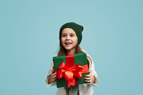 Happy little girl in casual outfit smiling and demonstrating wrapped present while standing against blue background - Zdjęcie, obraz