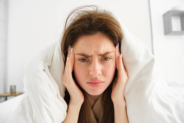 Woman feeling unwell in bed, lying in her bedroom under blanket and white sheets in morning, frowning and touching head, has headache or migraine. - Photo, Image