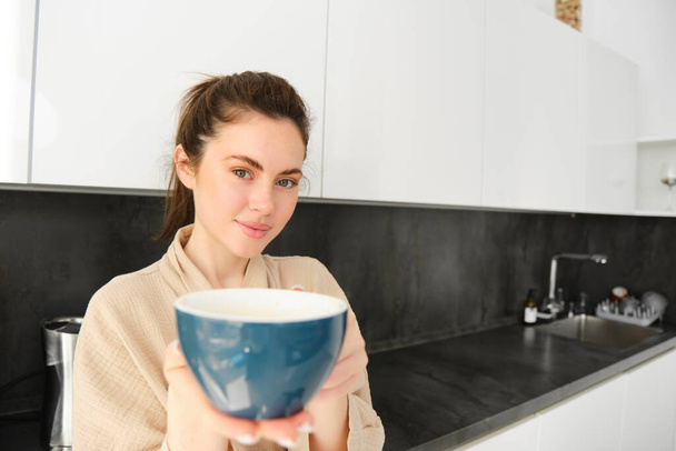 Portrait of good-looking young woman gives you cup of coffee, offering mug to you, standing in the kitchen, wearing bathrobe. - Photo, Image