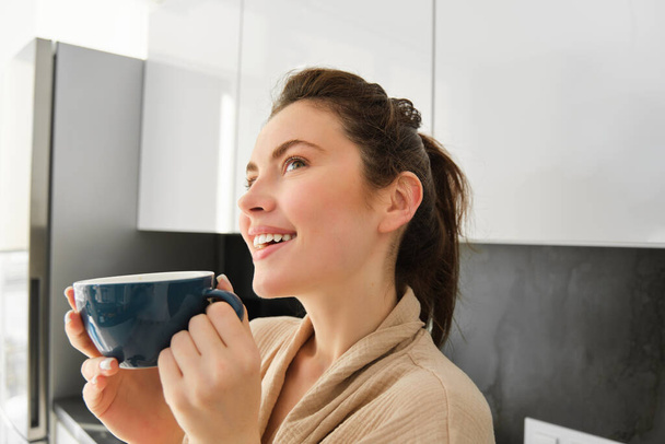 Közelkép portré álmodozó, mosolygós fiatal nő, iszik kávét, és úgy néz ki, figyelmes, áll a konyhában. - Fotó, kép