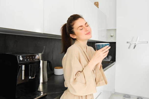 Portrét ženy stojící u kávovaru, připravující ranní šálek cappuccina, stojící v kuchyni, v županu. - Fotografie, Obrázek