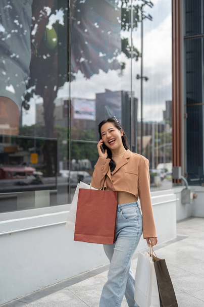 Привлекательная и веселая молодая азиатка в модной одежде разговаривает по телефону со своей подругой, наслаждаясь днем покупок в городе.. - Фото, изображение