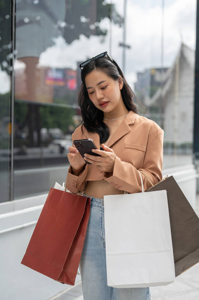 Moda kıyafetleri içinde muhteşem bir Asyalı kadın alışveriş çantasıyla şehirde yürürken akıllı telefonundan gelen mesajlara cevap veriyor. Alışveriş günü, Kentsel yaşam tarzı - Fotoğraf, Görsel