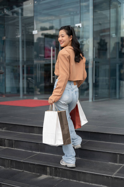 Una giovane donna asiatica gioiosa e bella in abiti alla moda sta entrando in un centro commerciale, camminando con le sue borse della spesa e godendo la sua furia commerciale in città nel fine settimana.. - Foto, immagini
