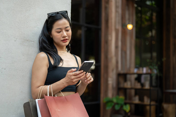 Una hermosa joven asiática en ropa de moda está respondiendo a los mensajes en su teléfono inteligente mientras disfruta de su día de compras en la ciudad. Día de compras, Estilo de vida urbano, Chica de ciudad - Foto, imagen