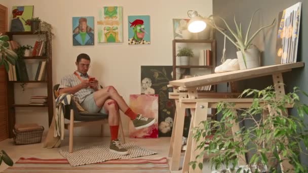 Ευρεία φωτογραφία του νεαρού καυκάσιου σχεδιαστή να κάθεται στην πολυθρόνα και να περιφέρεται στα μέσα κοινωνικής δικτύωσης σε smartphone στο στούντιο της σοφίτας με πίνακες στους τοίχους την ημέρα - Πλάνα, βίντεο