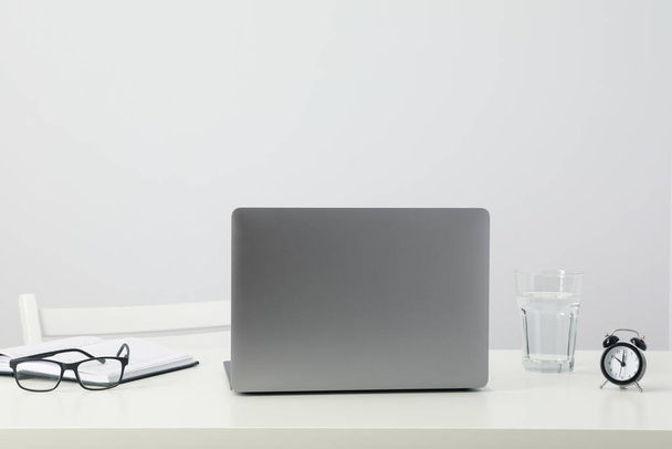 Ноутбук, очки, стекло и будильник на столе на белом фоне - Фото, изображение