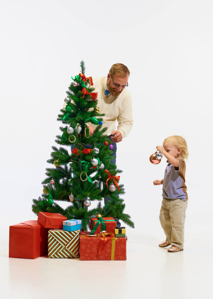 Boas férias de inverno. Criança feliz, menino com pai, homem decorando com brinquedos Árvore de Natal contra fundo de estúdio branco. Conceito de dezembro, Ano Novo, felicidade, infância. Espaço para cópia. ad - Foto, Imagem