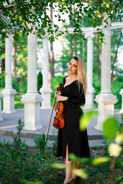 Beautiful blonde girl in black dress holding a violin in green park. Art work. Sad lady dreaming. - Photo, Image