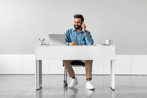 Homme d'affaires indien positif parlant sur smartphone, ayant une conversation, assis à table avec un ordinateur portable contre un mur léger, pleine longueur, espace de copie. Concept de communication à distance - Photo, image