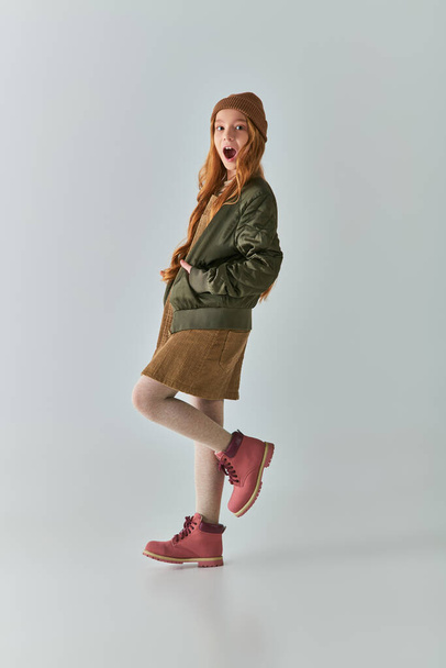 moda invernale, ragazza stupita con i capelli lunghi e cappello a maglia in piedi in abito e giacca su grigio - Foto, immagini