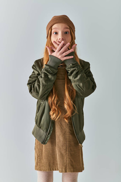 winter fashion, shocked girl with long hair and knitted hat standing in dress and jacket on grey - Foto, afbeelding