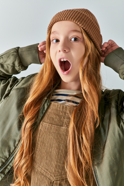 winter fashion, emotional girl with open mouth wearing hat and looking at camera on grey backdrop - Foto, Imagem