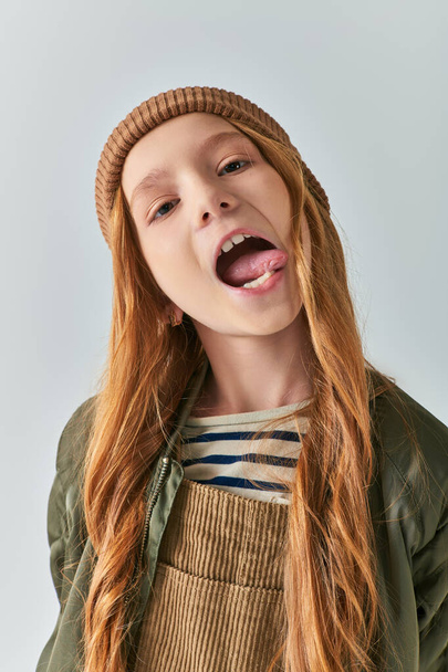 kiskorú lány kötött kalapban és felsőruházatban kilóg a nyelve, és a kamerát nézi szürke háttéren - Fotó, kép