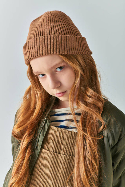 serious preteen girl in knitted hat and winter outerwear looking at camera on grey backdrop - Photo, Image