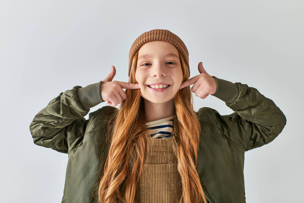 positive preteen girl in knitted hat and winter outfit pointing at cheeks with fingers on grey - Photo, Image