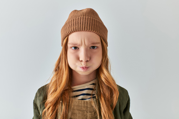 displeased preteen girl in stylish winter outfit with knitted hat puffing cheeks on grey background - Photo, Image
