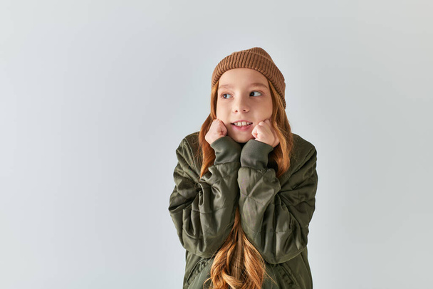 preteen girl in stylish winter outfit with knitted hat feeling cold while standing on grey backdrop - Fotografie, Obrázek