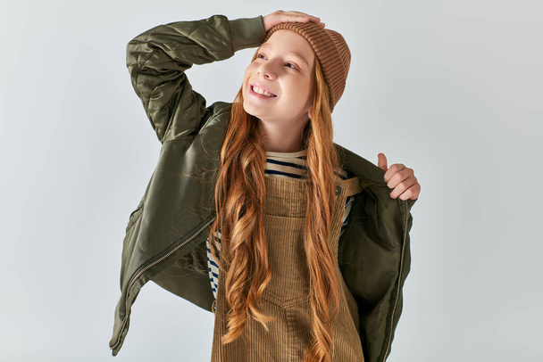 ευχαριστημένος preteen κορίτσι σε πλεκτό καπέλο και ζεστά ρούχα προσαρμογή σακάκι του χειμώνα σε γκρι φόντο - Φωτογραφία, εικόνα