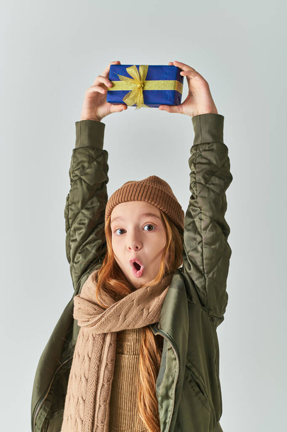 amazed girl in stylish winter outfit with hat holding Christmas present above head on grey backdrop - Valokuva, kuva