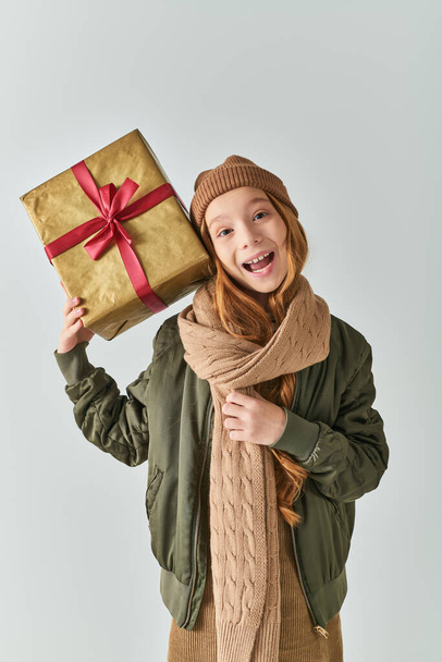 excited preteen girl in winter outfit with knitted hat holding Christmas present on grey backdrop - Photo, Image