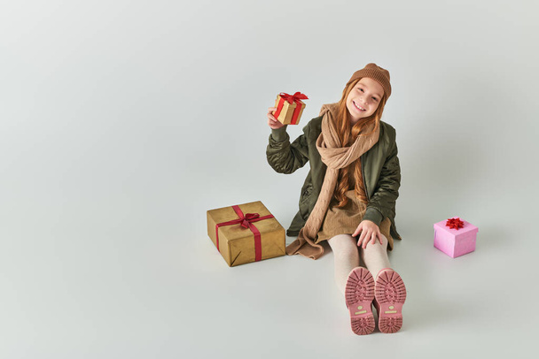 Şık kıyafetli, kışlık şapkalı, Noel hediyeli ve gri üzerine oturan mutlu ergen çocuk. - Fotoğraf, Görsel