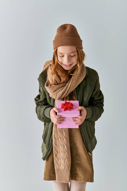 happy preteen kid in stylish outfit with winter hat holding Christmas present and standing on grey - 写真・画像