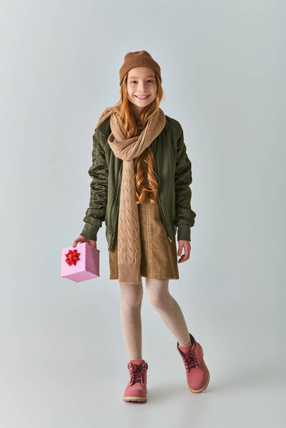 happy preteen girl in winter clothes and knitted hat holding Christmas present and standing on grey - Photo, Image