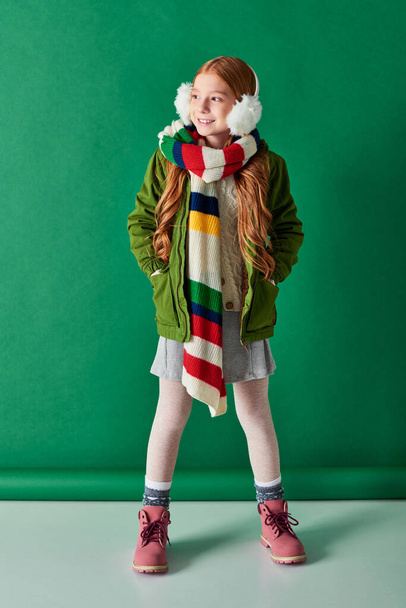 volledige lengte, vrolijk meisje in oorkappen, gestreepte sjaal en winter outfit op turquoise achtergrond - Foto, afbeelding