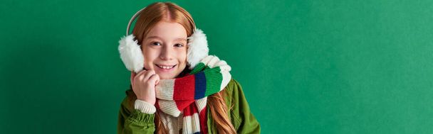 preteen girl in ear muffs, striped scarf and winter outfit smiling on turquoise backdrop, banner - Photo, Image