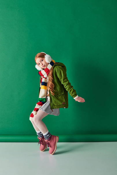 стильная девочка-подросток в наушниках, полосатый шарф и зимний наряд, позирующий на бирюзовом фоне - Фото, изображение