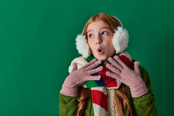 Şok olmuş kulaklıklı genç kız, çizgili atkı ve turkuaz arka planda el kol hareketi yapan kışlık kıyafet. - Fotoğraf, Görsel