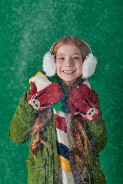cheerful girl in ear muffs, striped scarf and winter attire standing under falling snow on turquoise - Photo, Image