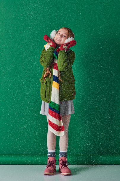 menina feliz em regalos de orelha, cachecol quente e traje de inverno de pé sob a neve caindo em turquesa - Foto, Imagem