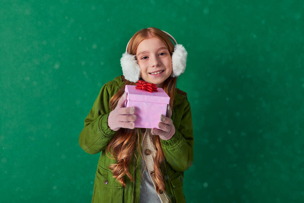 sezóna radosti, šťastný kluk v ušních manžetách a zimní oblečení držící sváteční dárek pod padajícím sněhem - Fotografie, Obrázek