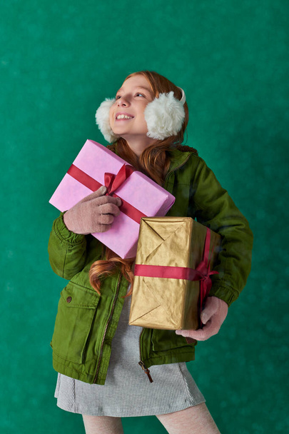 season of joy, pleased girl in winter outfit and ear muffs holding holiday gifts under falling snow - Photo, Image