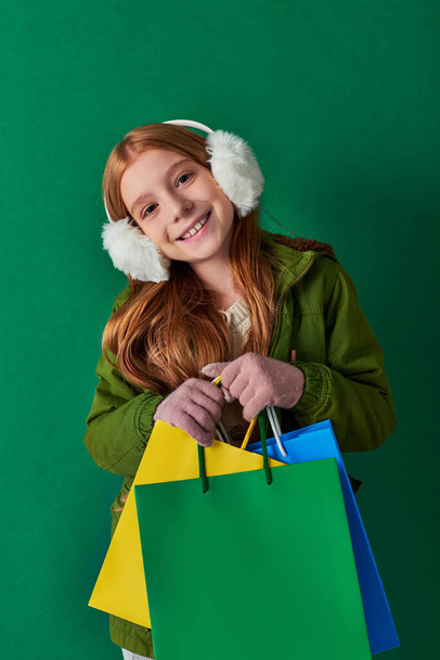 holiday season, joyful girl in winter outfit and ear muffs holding shopping bags on turquoise - Photo, Image