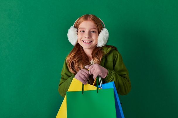 holiday season, excited kid in winter outfit and ear muffs holding shopping bags on turquoise - Photo, Image