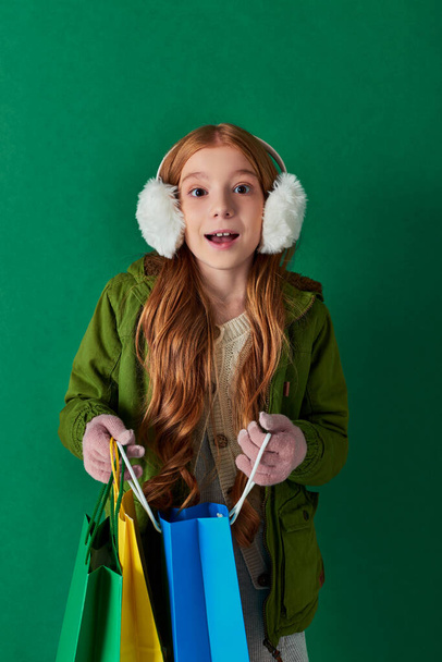зимние каникулы, возбужденный ребенок в зимнем наряде и наушники, держащие сумки для покупок на бирюзовом - Фото, изображение