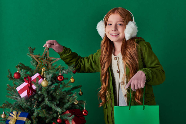winter holidays, happy girl in ear muffs holding shopping bag touching star top of Christmas tree - Photo, Image